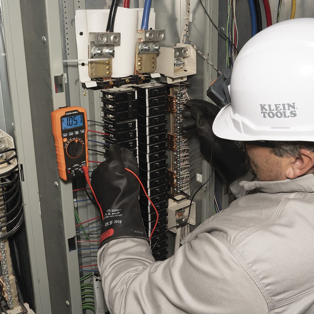 Action shot of a professional electrician using the Klein Tools MM420 Digital Multimeter to probe an industrial electrical cabinet. The technician is wearing high-voltage protective gloves while the multimeter display reads $105.1V$, showcasing the device's reliability for CAT III 600V safety environments.