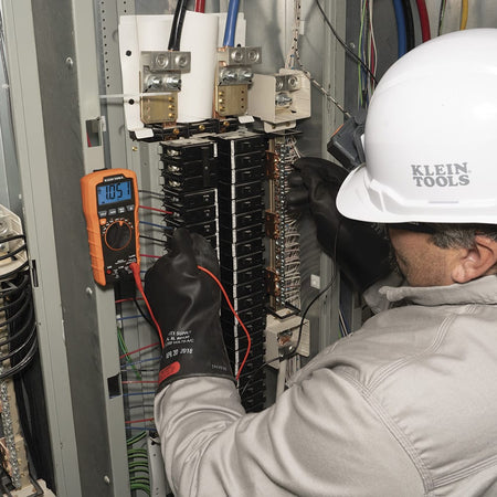 Action shot of a professional electrician using the Klein Tools MM420 Digital Multimeter to probe an industrial electrical cabinet. The technician is wearing high-voltage protective gloves while the multimeter display reads $105.1V$, showcasing the device's reliability for CAT III 600V safety environments.
