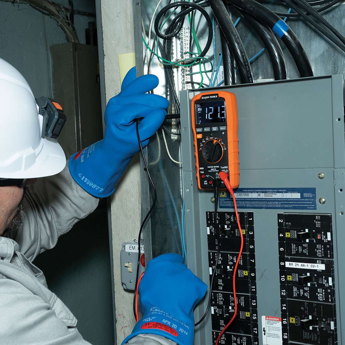An action shot showing a professional electrician using the MM720 to diagnose an industrial electrical panel. The technician, wearing insulated safety gloves and a hard hat, uses the test leads to verify a 121.3V reading on the backlit reverse contrast LCD, demonstrating the tool's performance in a commercial environment.