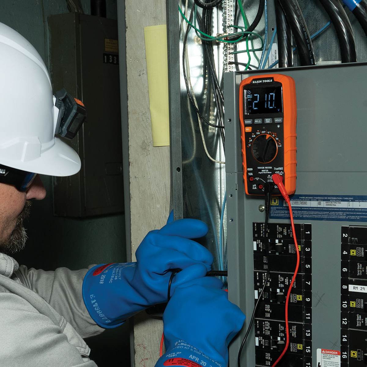 Person wearing a hard hat and blue gloves using an orange multimeter on electrical panel.