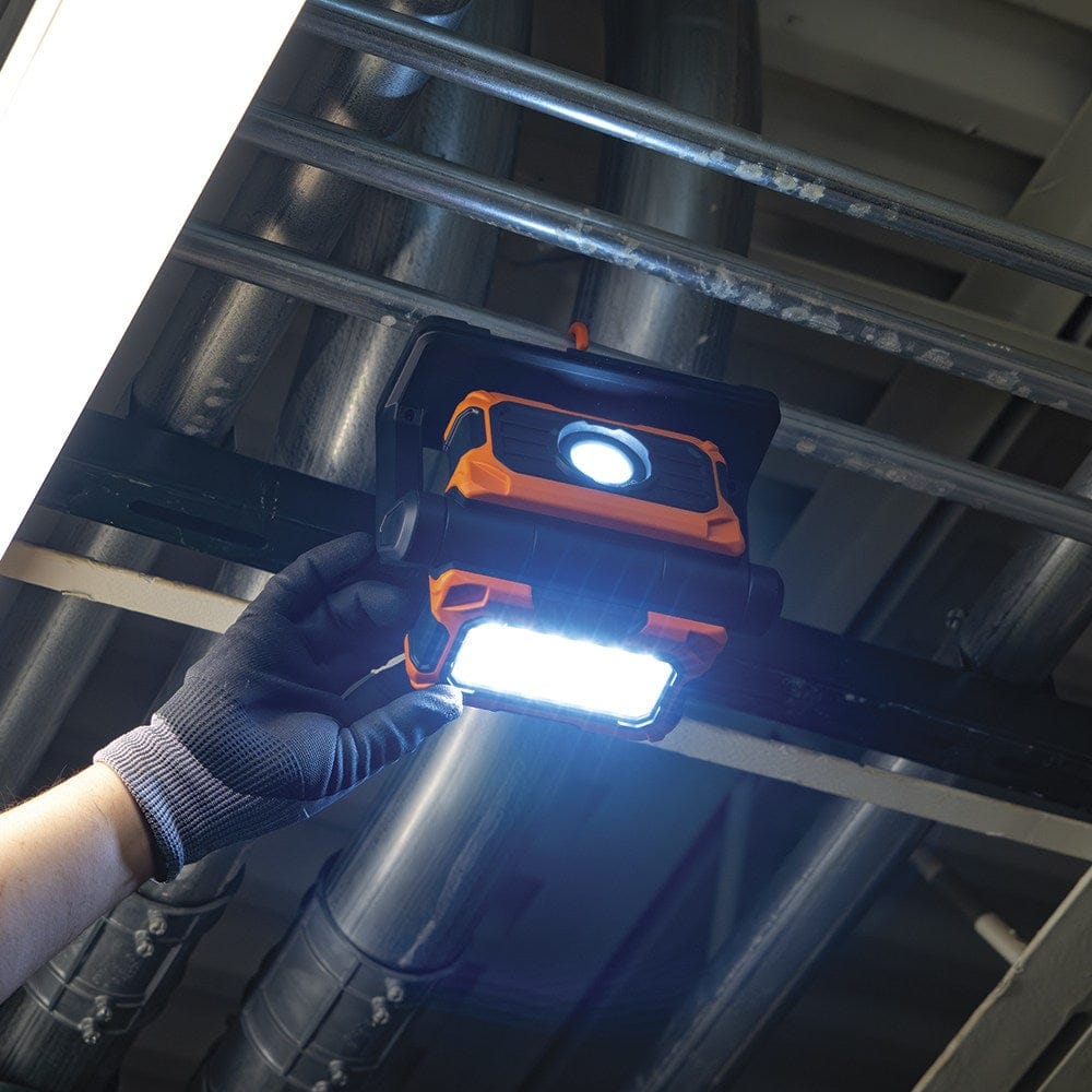 A professional tradesperson adjusting the rotating head of the Klein Tools 56417 while it is hung from overhead conduit pipes. The image demonstrates the ease of one-handed adjustment to direct the 3000-lumen beam in tight industrial ceilings.