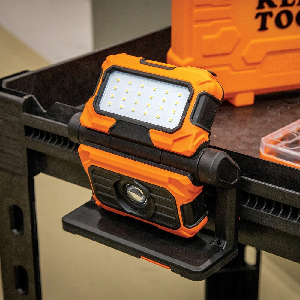 The Klein Tools rechargeable work light mounted to the edge of a black poly work table. The base is positioned to provide a stable platform while the flood light panel illuminates the workspace for detailed assembly or repair tasks.