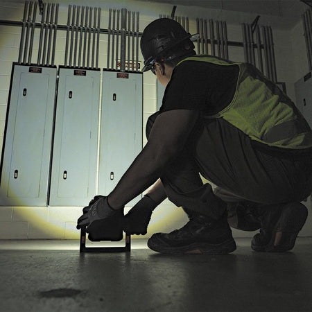 Low-angle shot of a technician in a hard hat crouching in a dark electrical room, using the Klein Tools 56417 on the floor to light up the bottom of a row of electrical panels. The image showcases the light's effective ground-level illumination.