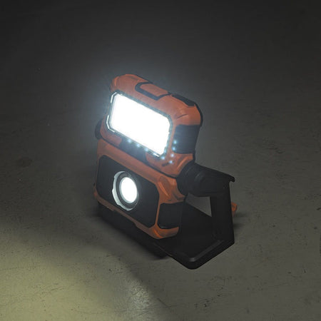 A studio shot of the rechargeable LED jobsite light standing on a concrete floor in a dark room. The light is tilted back on its rotating base, casting a bright white glow that illustrates its stability and ability to illuminate large floor spaces.