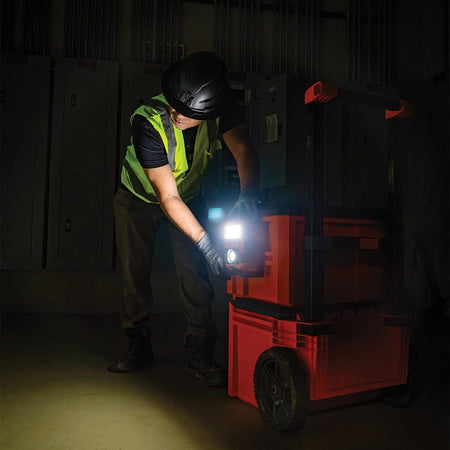 Action shot of a worker in a safety vest and hard hat attaching the Klein Tools work light to the side rail of a rolling MODbox tool stack. This highlights the seamless integration of the light into the MODbox mobile workstation system for easy transport.
