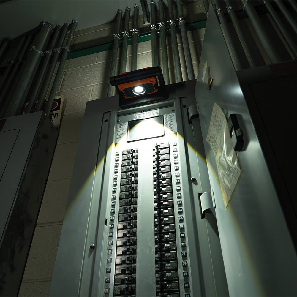 The magnetic base of the Klein Tools 56417 being used to mount the light onto a large grey electrical breaker panel. The spot light is directed downward, providing hands-free illumination for an electrician performing maintenance or inspections.