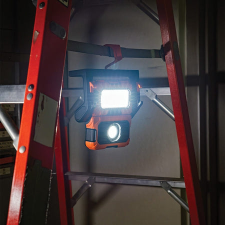 The integrated retractable hook of the Klein Tools work light is used to hang the unit from a red step ladder. This setup provides elevated, overhead flood lighting for projects where magnetic surfaces or floor space are unavailable.