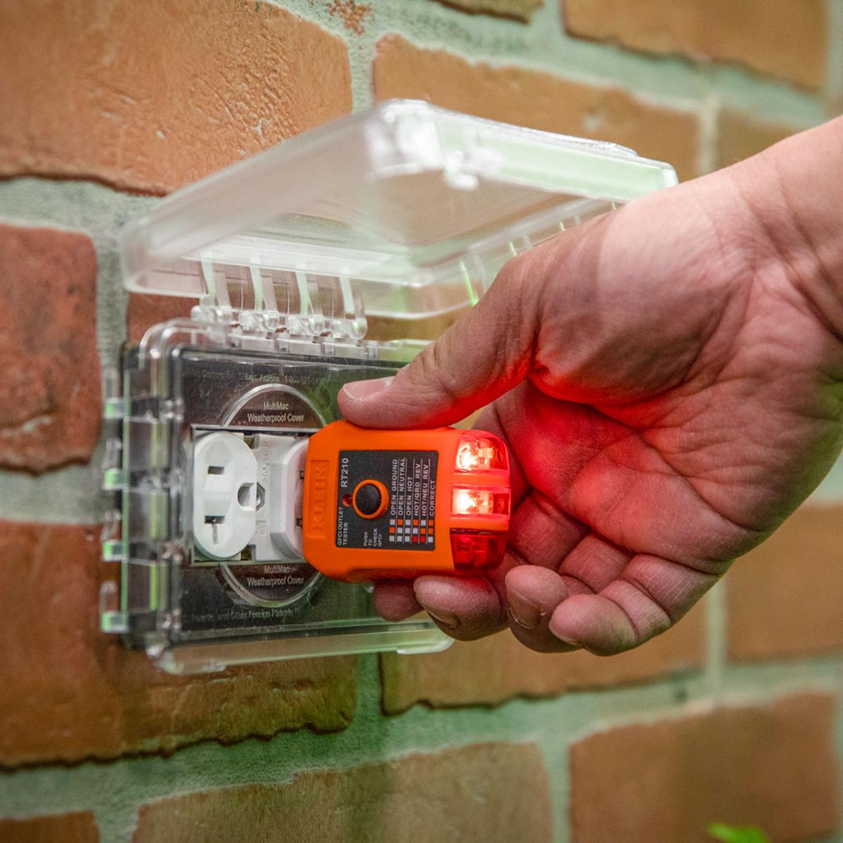 Action shot of a technician using the RT210 GFCI tester on an outdoor receptacle protected by a weatherproof cover. Two red LEDs are illuminated, signifying a correct wiring condition and active power in an exterior residential setting.