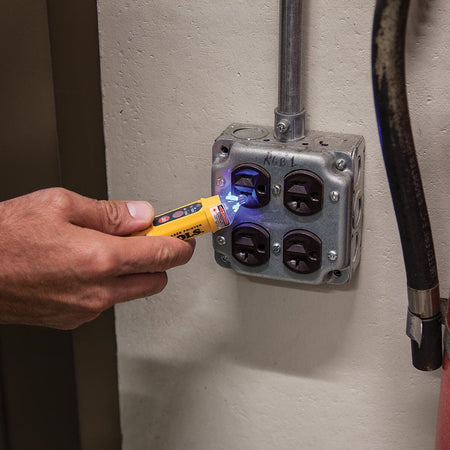 Action shot showing a technician using the NCVT-5A non-contact voltage tester to verify power at a commercial electrical junction box. The integrated LED indicator is illuminated with a bright blue light, demonstrating the tool's effectiveness in residential or industrial troubleshooting.