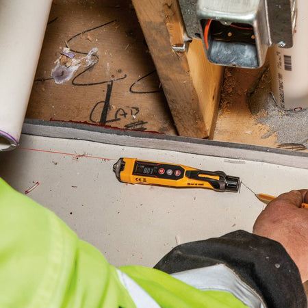High-angle action shot of the Klein Tools NCVT6 being used on a construction jobsite to mark a specific distance. The integrated laser projects a bright red line across a concrete surface while a worker uses a pencil to mark the measurement, showcasing the tool's utility in rough-in electrical layouts.
