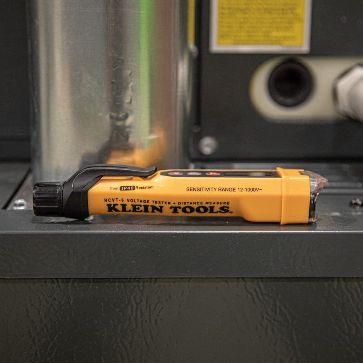 A side profile of the NCVT6 voltage tester and laser distance meter resting on a gray industrial electrical cabinet. The image highlights the "Sensitivity Range 12-1000V" branding and the IP40 dust-resistant rating, emphasizing its rugged design for professional environments.