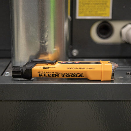 A side profile of the NCVT6 voltage tester and laser distance meter resting on a gray industrial electrical cabinet. The image highlights the "Sensitivity Range 12-1000V" branding and the IP40 dust-resistant rating, emphasizing its rugged design for professional environments.