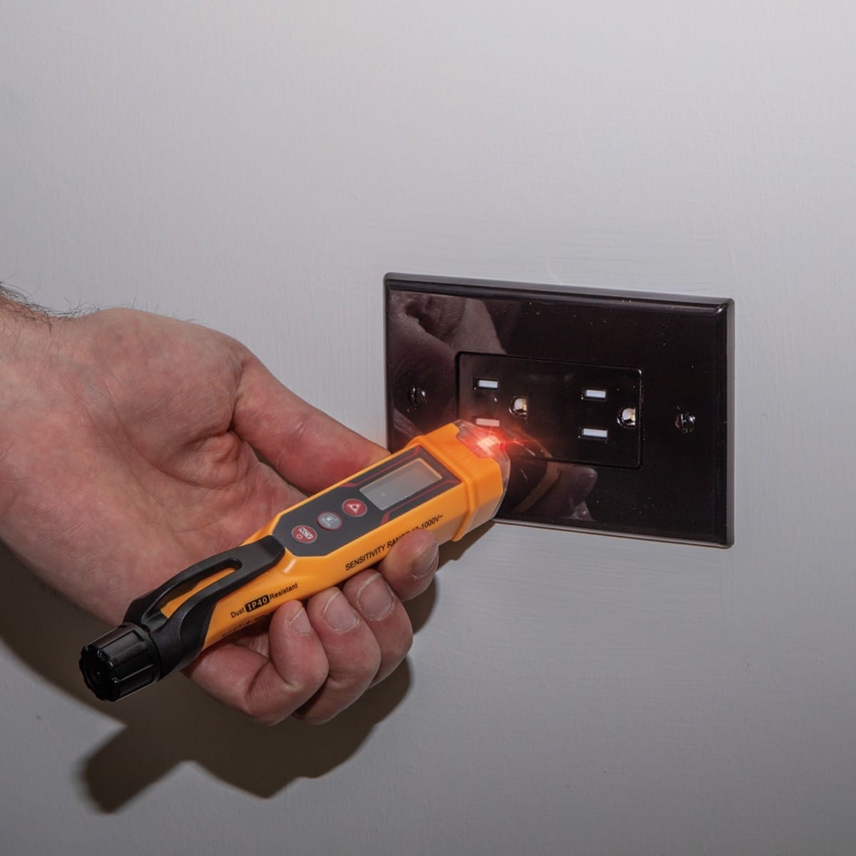 A technician using the Klein Tools NCVT6 to check a standard black electrical wall outlet. The clear tip is illuminated with a red LED, providing a simultaneous visual alarm that live voltage is present in the circuit.