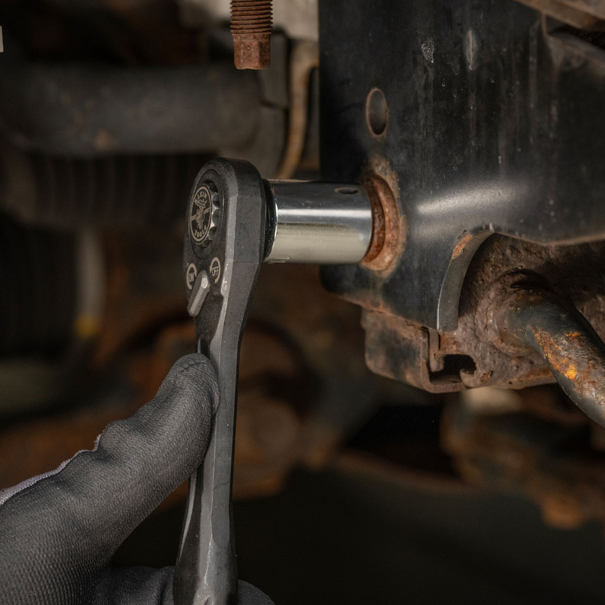 Mechanic using the low-profile Klein pass-through ratchet and a standard socket adapter to loosen a tight chassis bolt in a cramped bay.