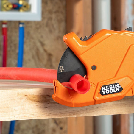 Klein Tools precision ratcheting PVC pipe cutter 50044 shown cutting through a red PEX pipe on a wooden workbench. The heavy-duty steel blade is engaged in a clean, straight cut, demonstrating the tool's precision and 1-1/4 inch cutting capacity.