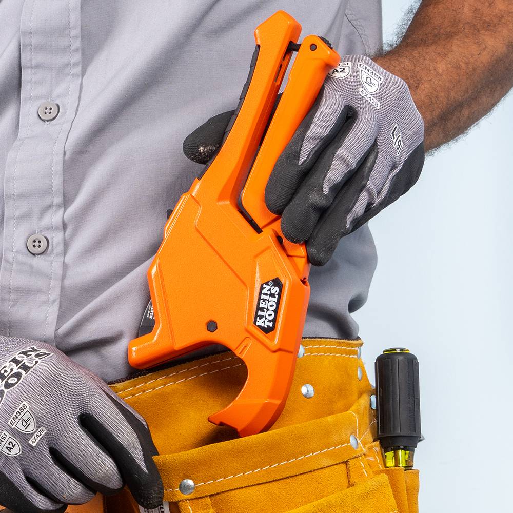 A professional tradesperson wearing protective gloves stores the orange Klein Tools ratcheting pipe cutter in a leather tool belt. The image highlights the compact, ergonomic design and portability of the 50044 PEX and CPVC cutting tool for job site efficiency.
