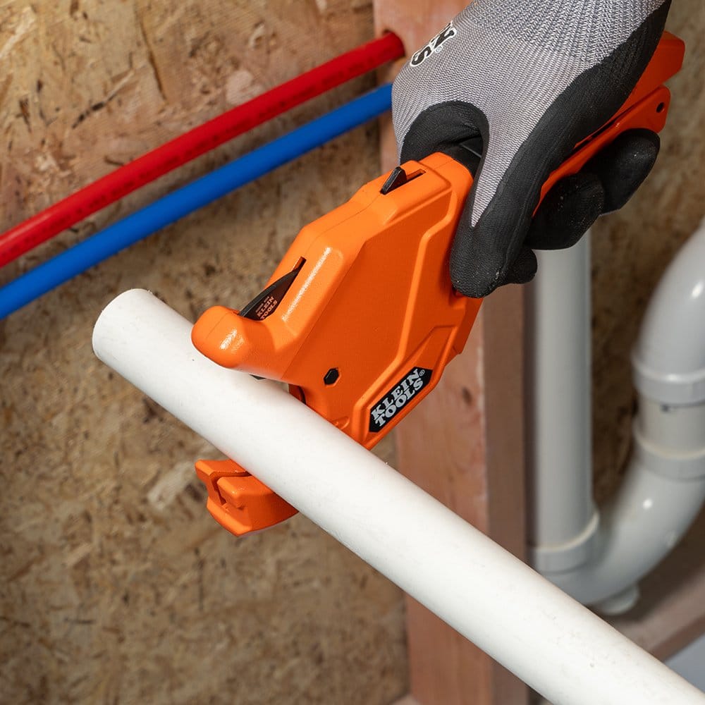 Top-down view of a plumber using the Klein Tools 50044 ratcheting cutter to slice through a white PVC pipe. The comfort grip handle and ratcheting mechanism are visible, showcasing how the tool provides maximum leverage for effortless heavy-duty plumbing cuts.