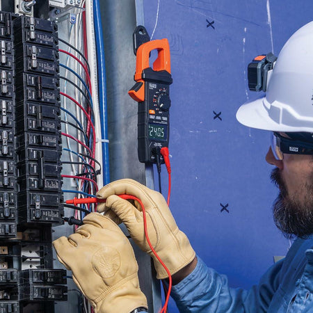 Person wearing safety gear using Klein Tools clamp meter to measure voltage in a circuit breaker panel; display reads 240.5 volts.