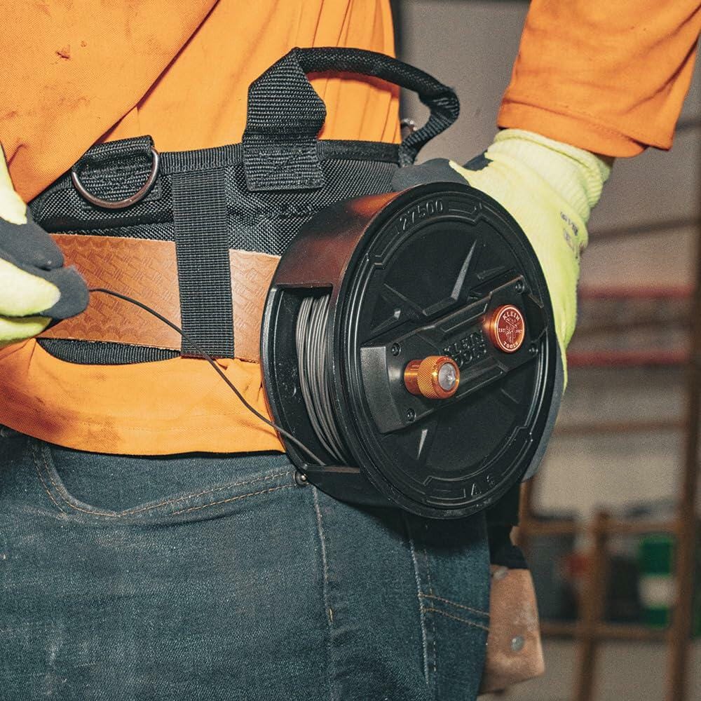 Close-up of the Klein Tools Tie Wire Reel attached to a worker's tool belt, showing a roll of wire installed and the spool mechanism secured by the quick-lock lid.