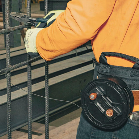 A construction worker is seen from the side, wearing the Tie Wire Reel and using pliers to twist tie wire onto a rebar cage.