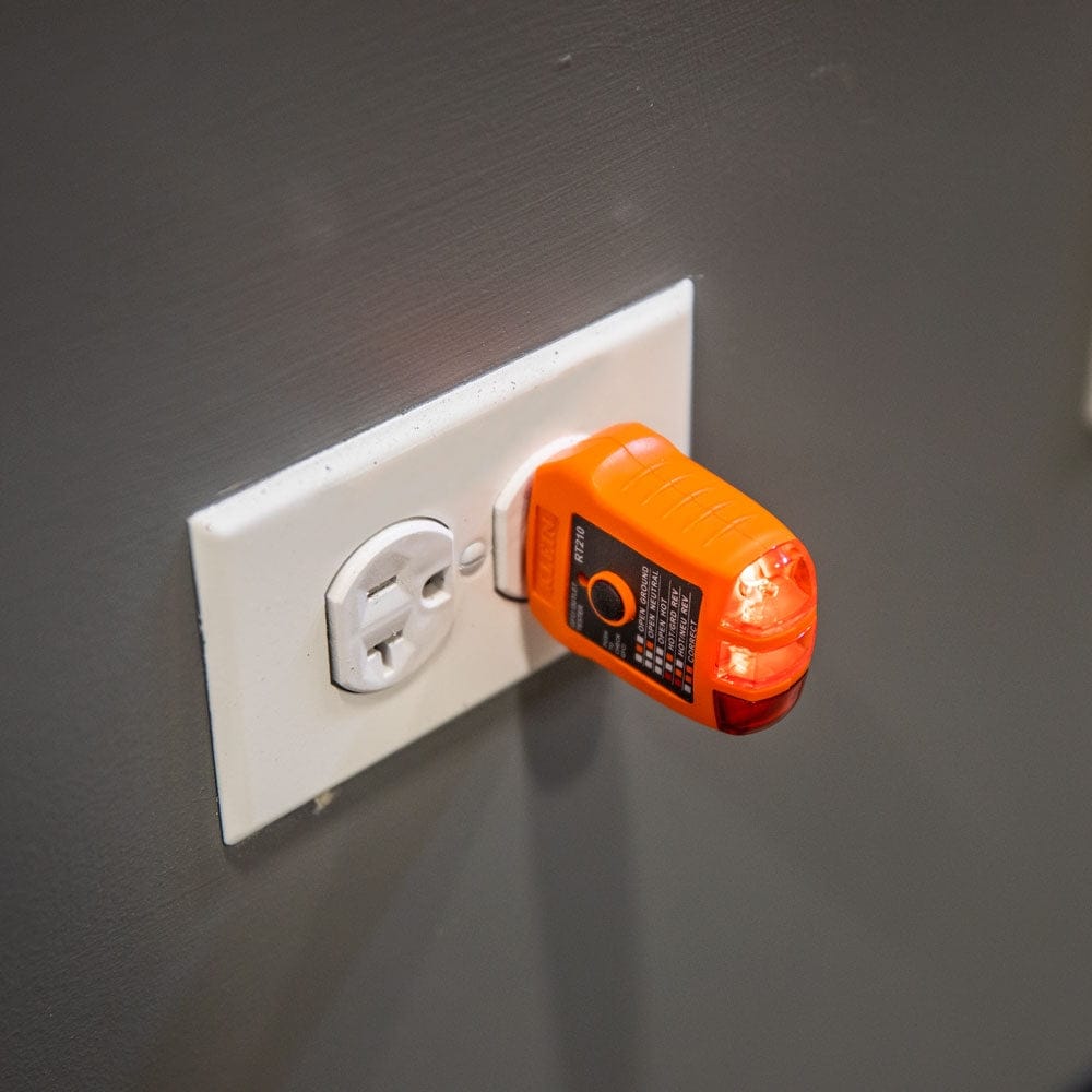 Action shot showing the RT210 tester plugged into a white wall outlet; two orange lights are illuminated on the end of the tool to indicate the wiring condition.