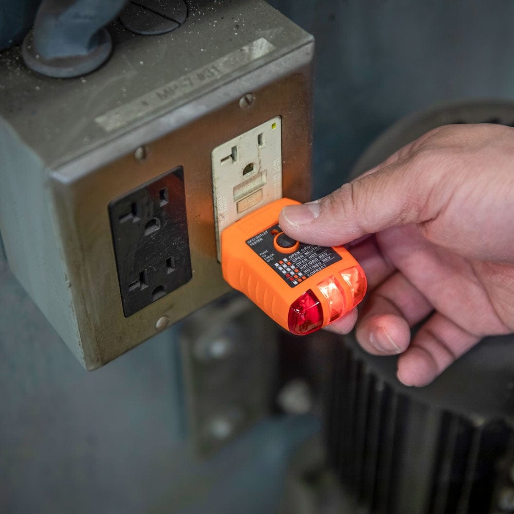 A close-up action shot showing a person's thumb pressing the black GFCI test button while the orange tester is plugged into a professional-grade metallic electrical box.