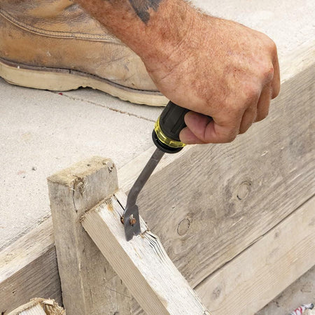 Close-up of a construction worker using a black and yellow flat pry bar to remove a rusted nail from wood, with tan work boots and tattooed forearm visible on a concrete surface.