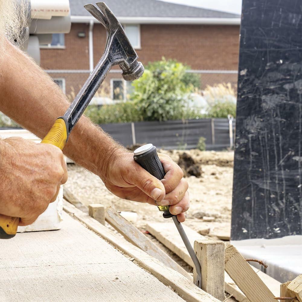Close-up of construction worker’s hands using hammer and screwdriver to adjust wooden form on concrete surface at outdoor site, with residential building in background.