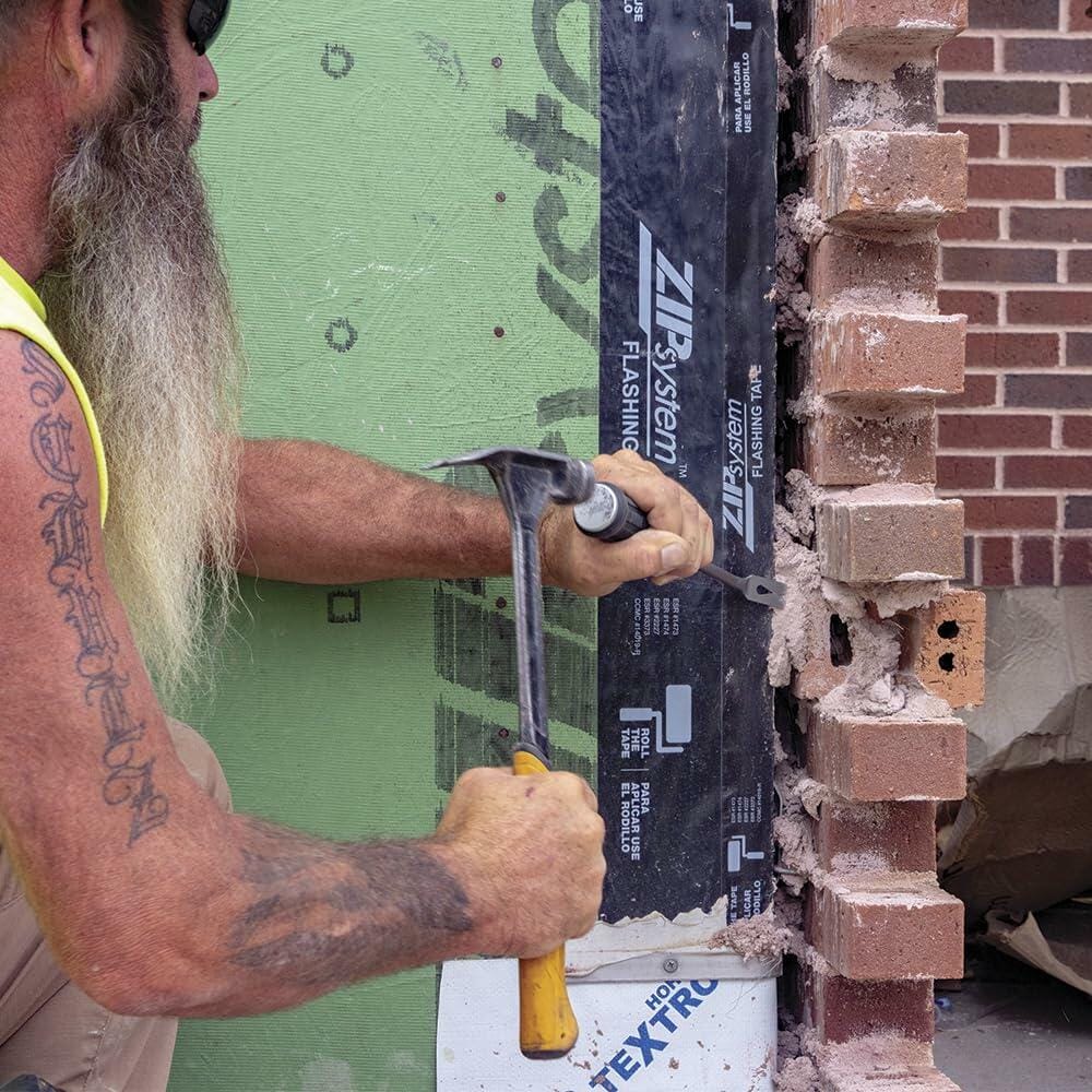Worker with long beard and tattoos using hammer and chisel to remove red bricks from wall with green ZIP System sheathing and black flashing tape, wearing sunglasses and yellow sleeveless shirt.