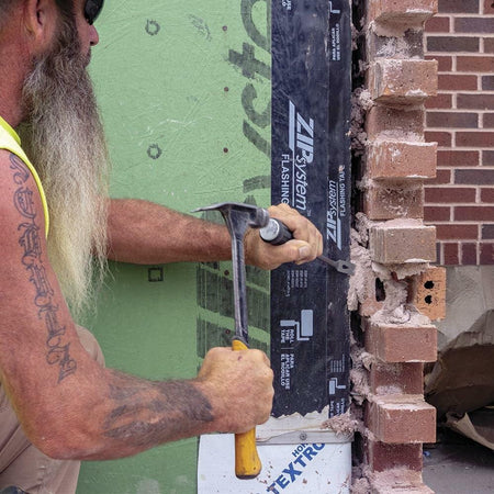 Worker with long beard and tattoos using hammer and chisel to remove red bricks from wall with green ZIP System sheathing and black flashing tape, wearing sunglasses and yellow sleeveless shirt.