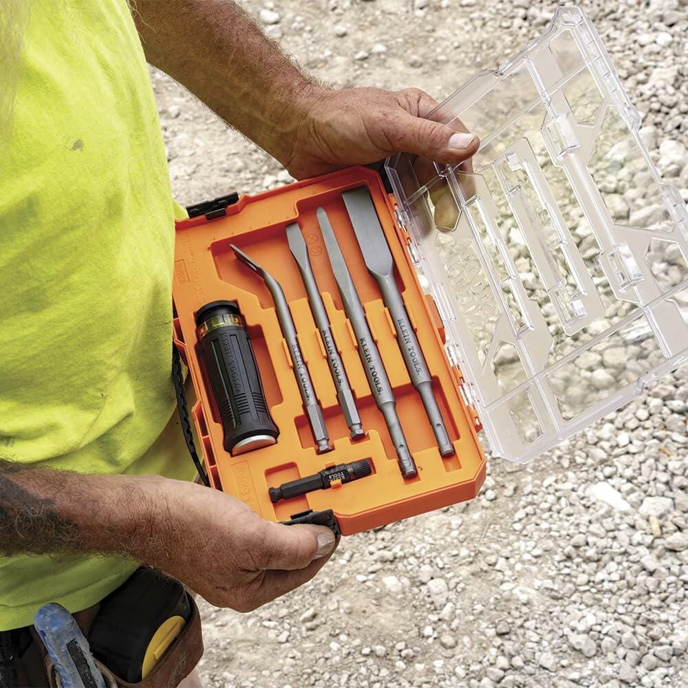 Open plastic tool case with bright orange interior showing five chisels, a black and yellow handle, and accessories, held by a worker wearing a yellow shirt and tool belt at a gravel construction site