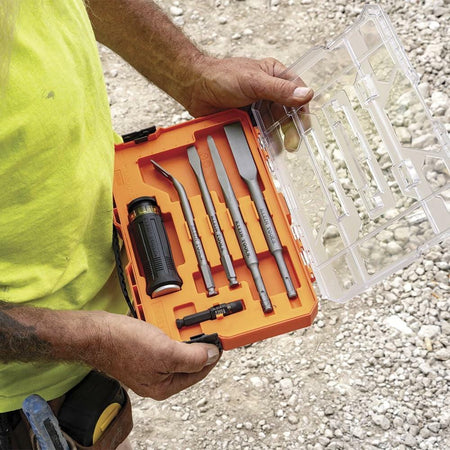 Open plastic tool case with bright orange interior showing five chisels, a black and yellow handle, and accessories, held by a worker wearing a yellow shirt and tool belt at a gravel construction site