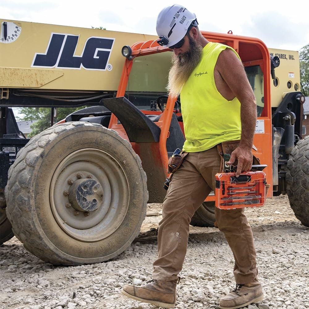 Construction worker in yellow sleeveless shirt and white hard hat walking on gravel site carrying orange toolbox, with JLG telehandler and tool belt visible in the background.