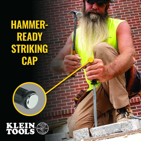 Worker kneeling on one knee using a hammer and labeled KLEIN TOOL CO. chisel on a concrete block, wearing tan pants, work boots, and yellow sleeveless shirt in front of a brick wall.