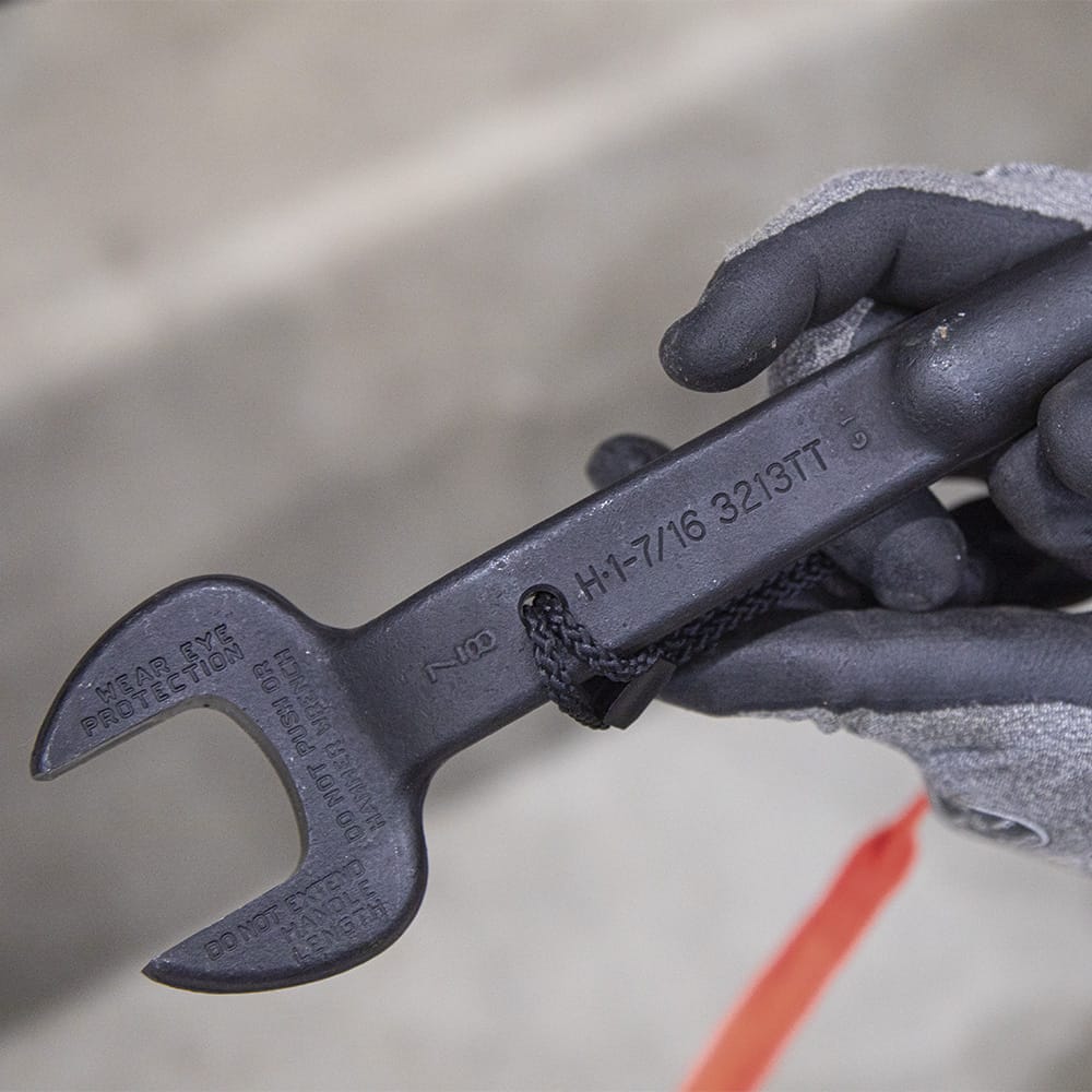 Gloved hand holding the head of a Klein 3213TT spud wrench displaying stamped 1-7/16-inch opening and 7/8-inch bolt size identification marks.