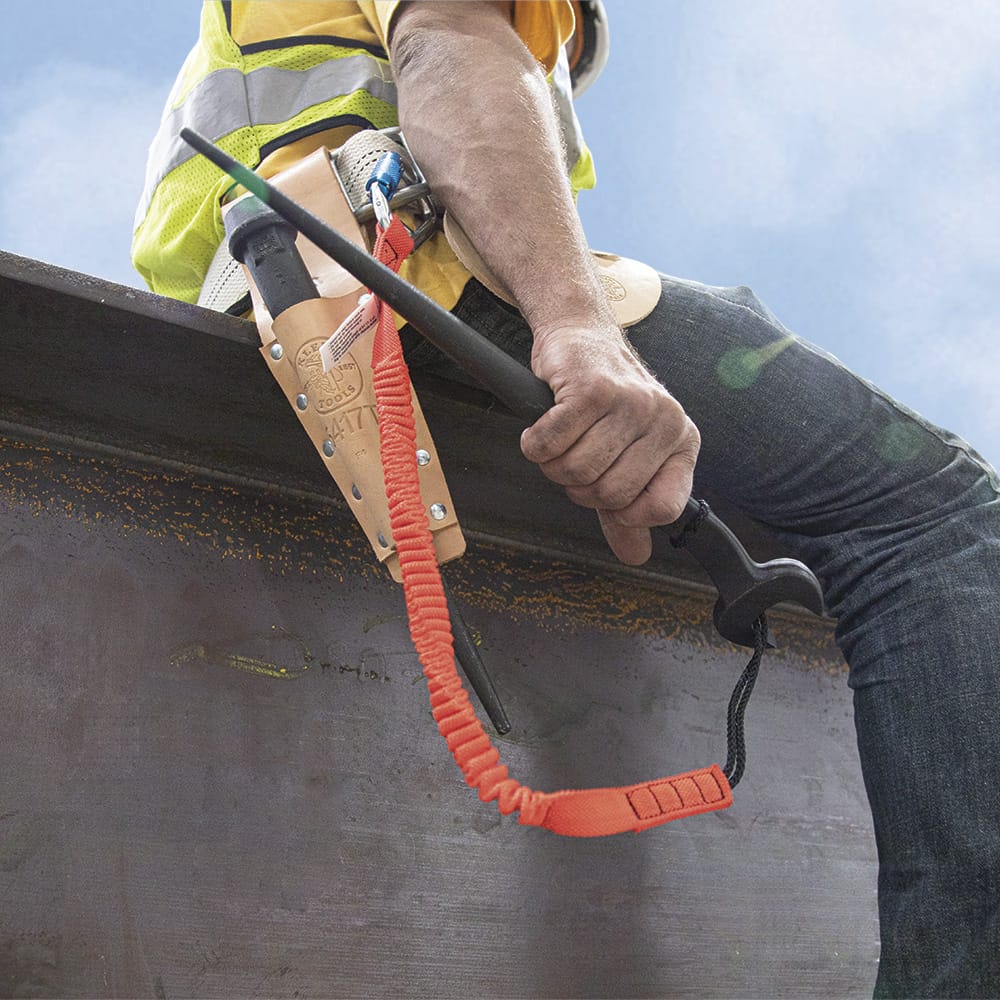 Ironworker sitting on a structural steel I-beam holding a tethered Klein 3213TT spud wrench connected to an orange fall-protection safety strap.