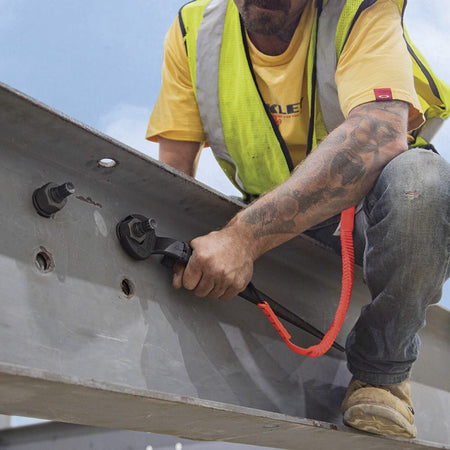Ironworker crouching on an I-beam using a tethered Klein 3214TT offset spud wrench to tighten heavy bolts on structural steel joints.