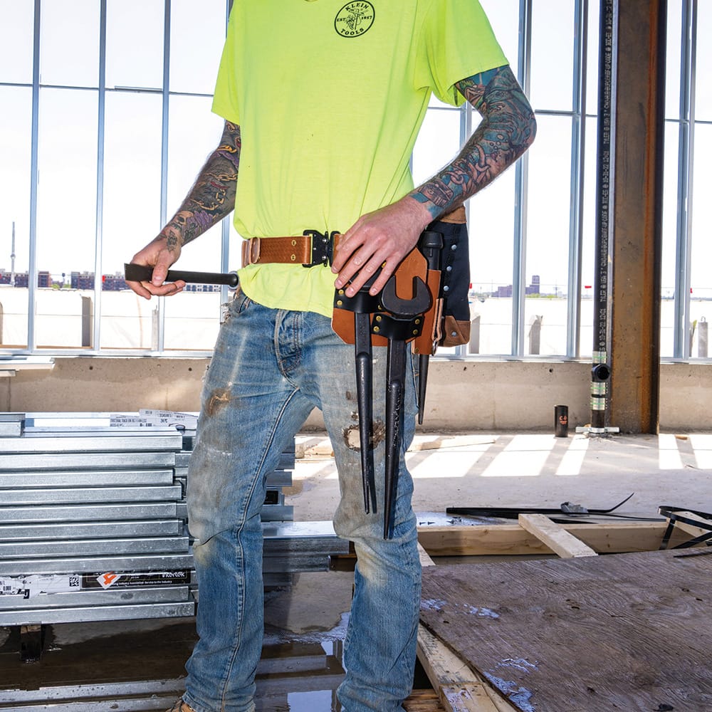 Ironworker standing on a construction floor showing the Klein 5860 leather holster mounted closely against a 3-inch tool belt via the tucked tunnel loop.