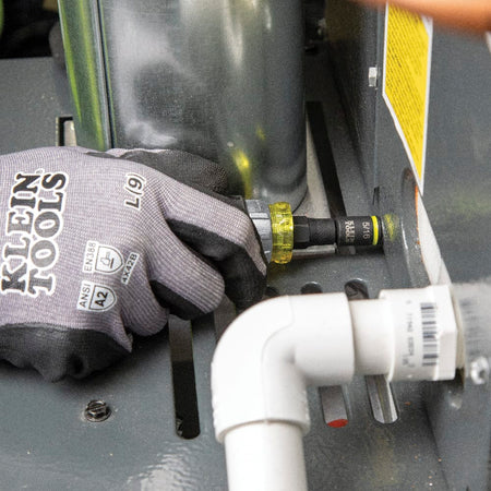 Worker maneuvering the Klein Tools 32561HDRT stubby driver in a cramped space behind PVC piping to adjust an HVAC panel fastener.