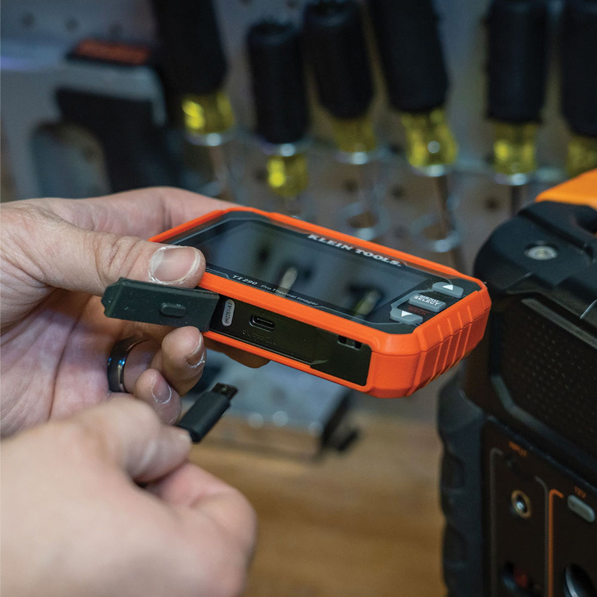 Person holding an orange and black device with 'Klein Tools' branding in a workshop setting.