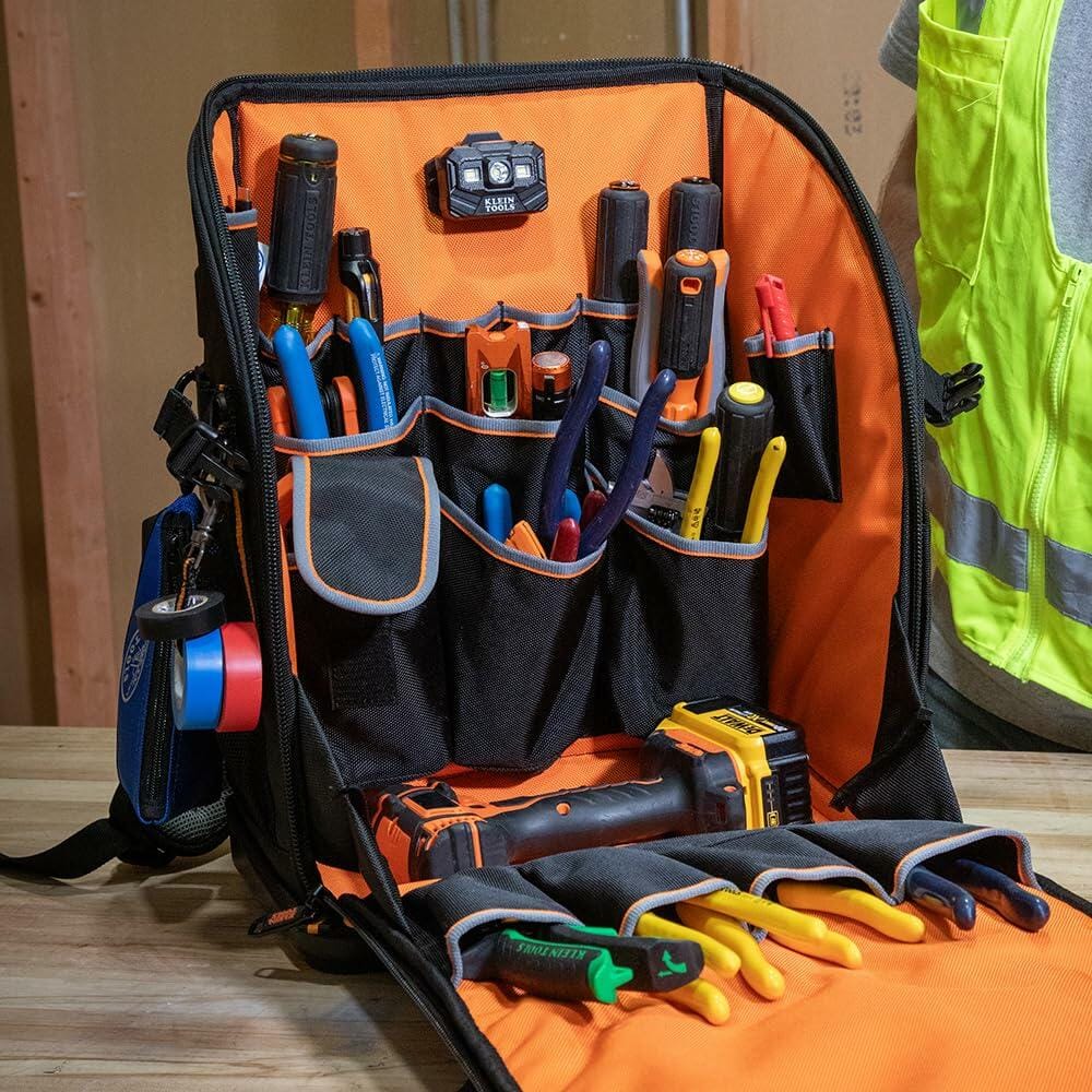 The backpack fully open and functioning as a workstation, displaying an organized array of tools in the pockets and a power tool lying in the bottom bay. A headlamp is mounted internally.