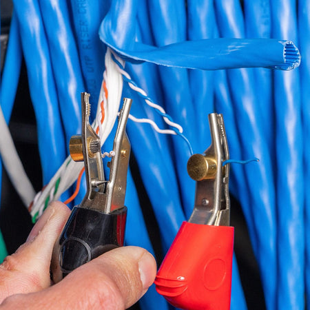 A detailed close-up of the rugged angled bed of nails (ABN) clips from the VDV500-820 kit. The clips are shown securely biting into the conductors of a blue CAT6 data cable, demonstrating how the specialized pins allow for precise electrical connection and toning without stripping the wire insulation.