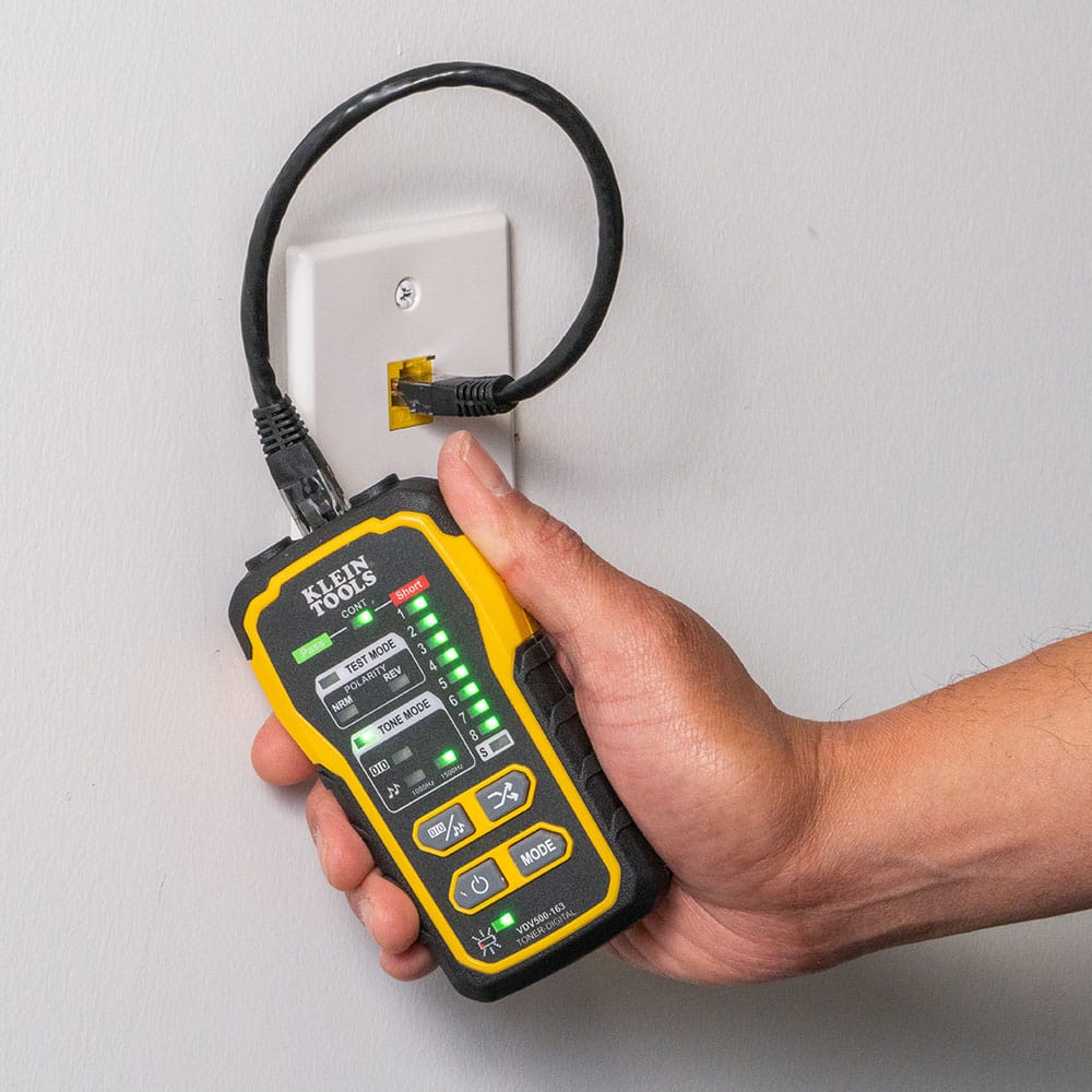 Action shot of a professional technician using the VDV500-163 digital tone generator to verify a network run. The unit is connected to an RJ45 wall plate with a patch cable, showing the diagnostic LED sequence used to confirm continuity, polarity, and pin-to-pin mapping on an active data network.