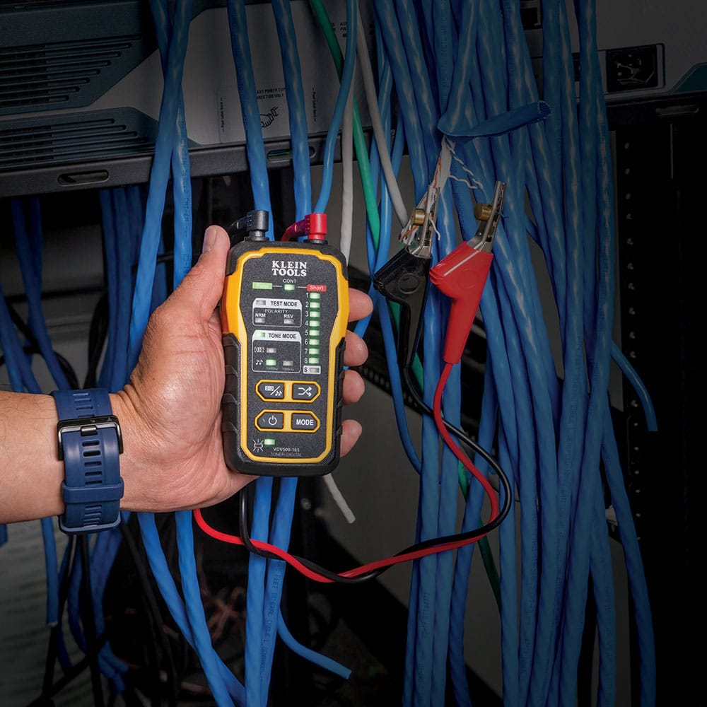 Action shot of a technician using the VDV500163 tone generator to test a data cable run. The generator is shown with its interactive LED display active, while the angled bed of nails clips are securely connected to an unterminated wire pair for active network tracing.