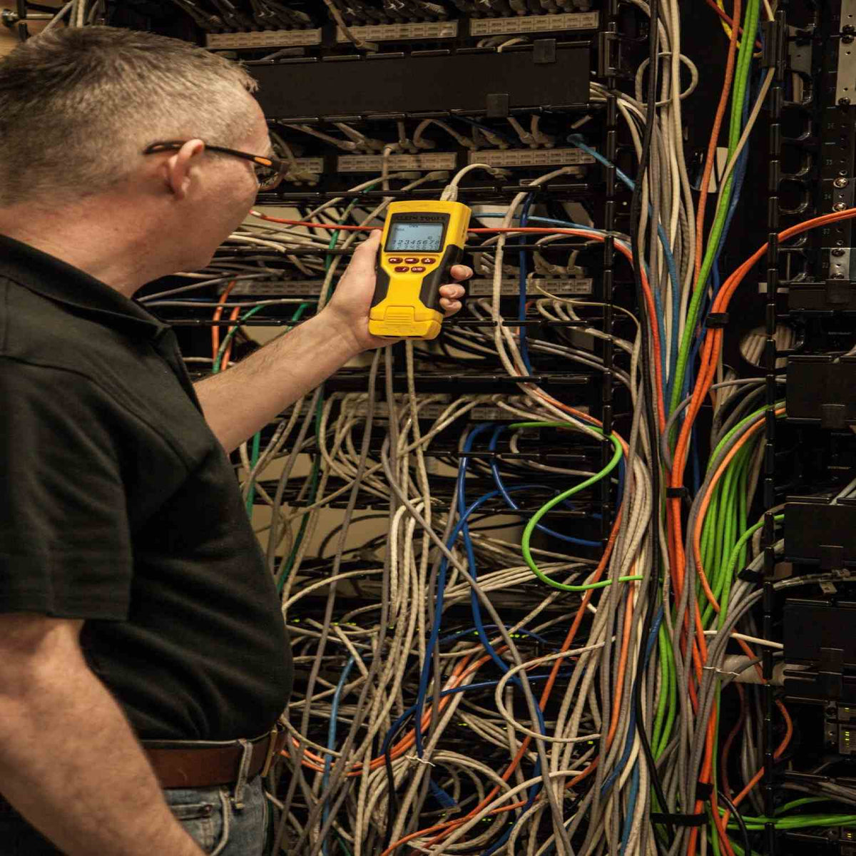 An in-the-field usage photo showing a network administrator in a complex server rack environment using the Klein Tools VDV501-825 Scout Pro 2 LT VDV tester to perform a wire map and continuity test on a specific patch panel port.