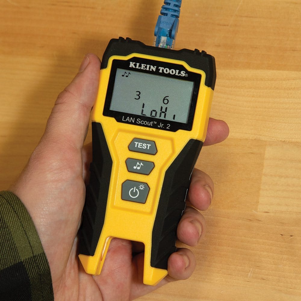 A close-up of the cable tester being held by a user, showing the device in tone mode with a musical note icon and the screen displaying LoH frequency indicators while connected to a blue patch cable.