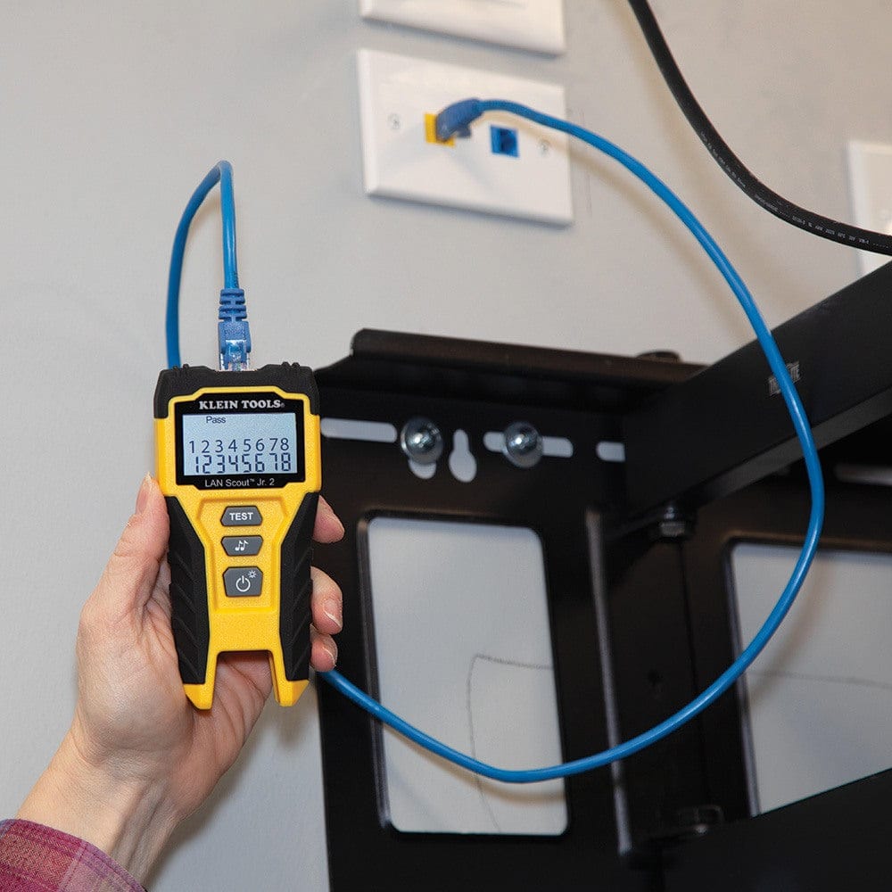 The tester plugged into a blue patch cable connected to a network wall jack, showing a successful Pass result on the backlit display.