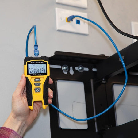 The tester plugged into a blue patch cable connected to a network wall jack, showing a successful Pass result on the backlit display.