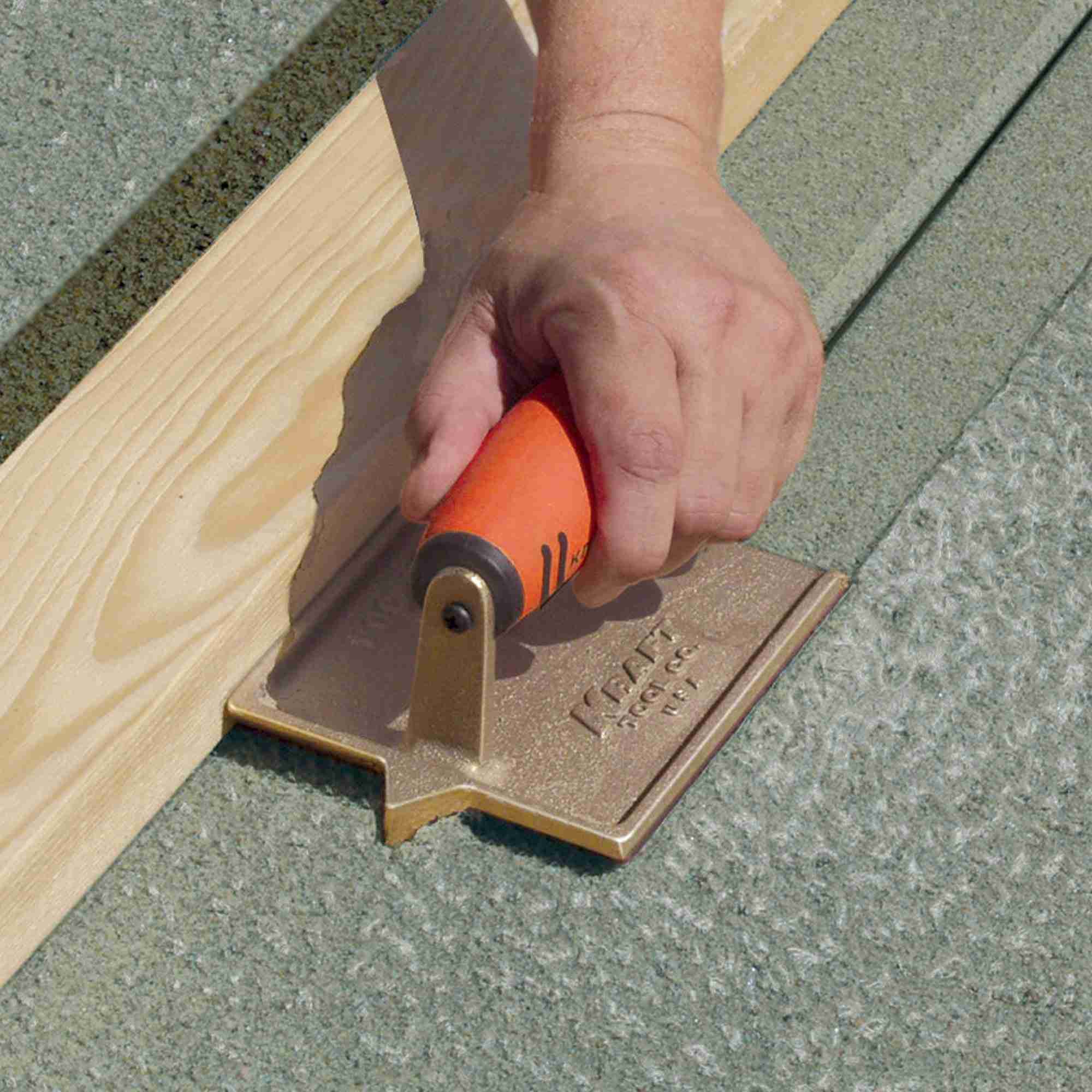 Close up action shot of a worker using the bronze hand groover to cut a joint along a wooden form board in wet concrete.