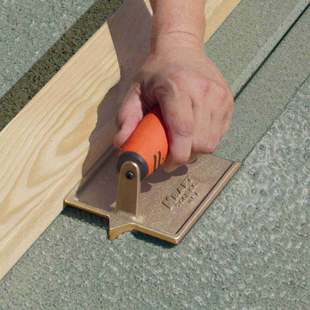 Close up action shot of a worker using the bronze hand groover to cut a joint along a wooden form board in wet concrete.